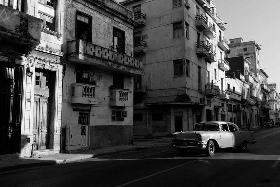 Shadowy black-and-white street scene in Havana with a 1950s vintage car passing weathered apartment buildings.