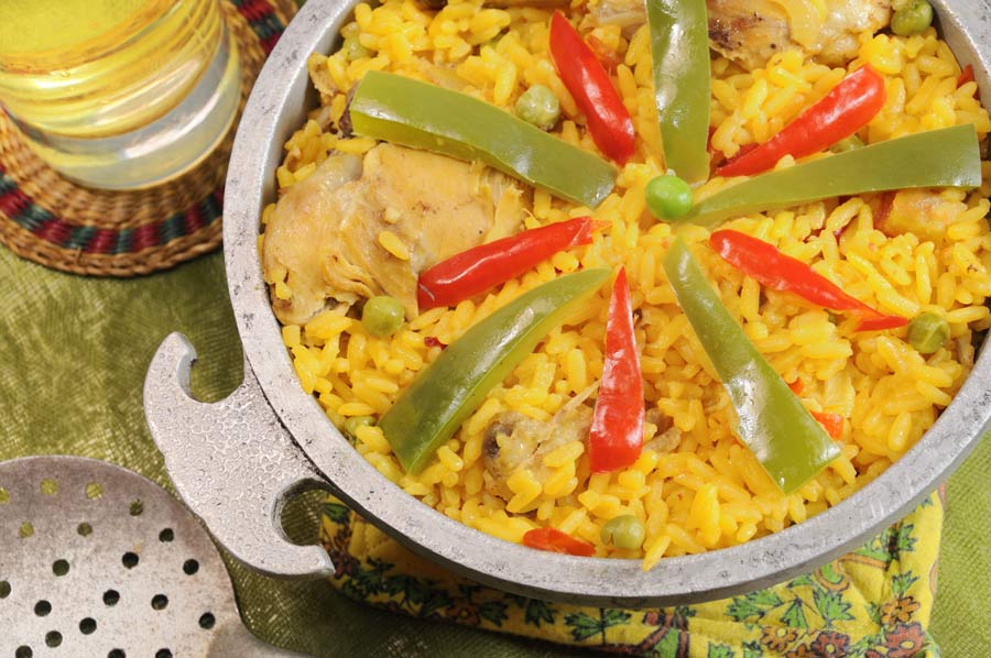 Traditional rice and chicken dish, arroz con pollo, topped with sliced red and green peppers, served in a metal pan on a colorful tablecloth along a serving ladle and a drink.