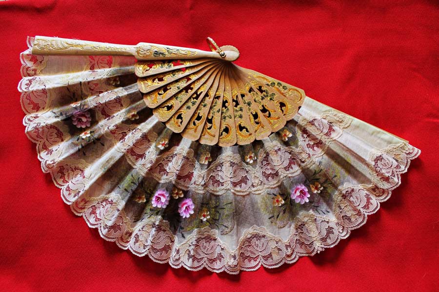 Decorative folding hand fan with floral lace and a carved wooden frame displayed on a rich red fabric background.