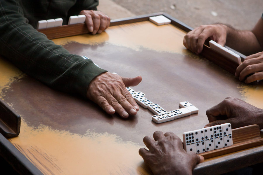 A close-up image showing only the hands of three people playing dominoes around a wooden table, reaching toward the black-and-white tiles during the game.