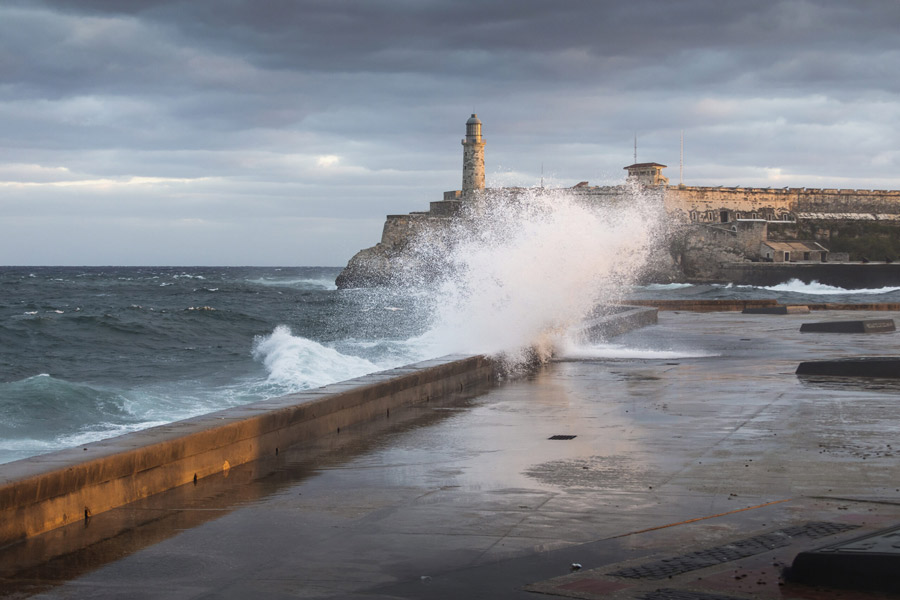 Waves crash against Havana’s seaside wall, sending spray into the air beneath a cloudy sky with El Morro Castle and its lighthouse in the distance.
