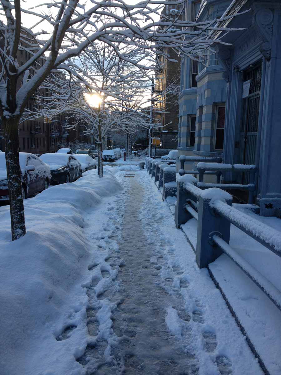 Snow-covered New York City sidewalk lined with buildings, parked cars, and bare trees at sunset, with footprints in the snow.