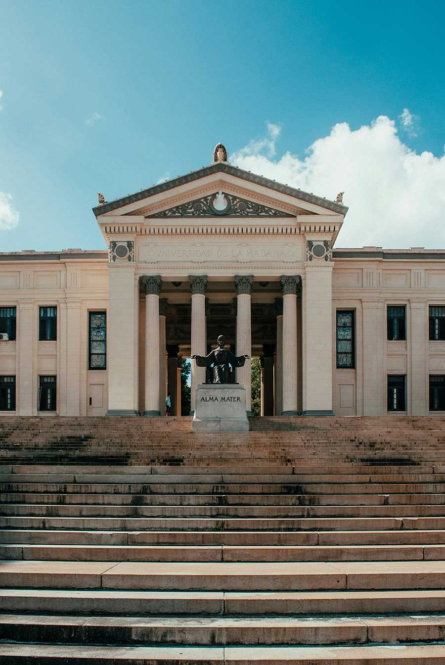 Grand neoclassical building of University of Havana viewed from the base of the wide stone steps on a sunny day with the Alma Mater statue at the top.